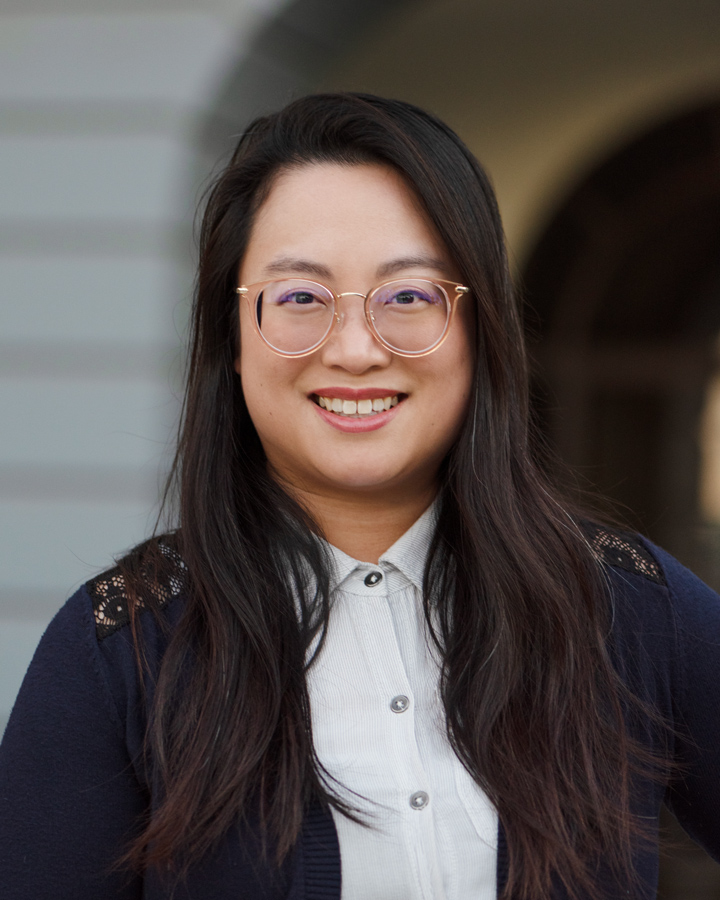Headshot of Christine, founder and proprietor of Say Front. She has long dark hair and is smiling warmly at the camera while wearing round gold-rimmed glasses. Christine is dressed professionally in a white button-up shirt with a navy cardigan featuring lace detailing at the shoulders. The image is taken against a soft-focus architectural background with an archway, creating a welcoming yet professional atmosphere.